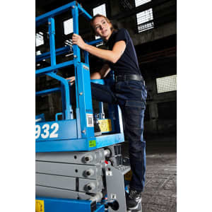 A woman in a black top and navy stretch jeans climbs a blue lift in a warehouse setting.