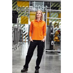A woman stands wearing an orange long-sleeve polo shirt and black workwear cargo pants in a store environment.