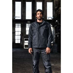A man wearing dark grey workwear pants and a matching jacket stands in an industrial setting. The image features a logo.