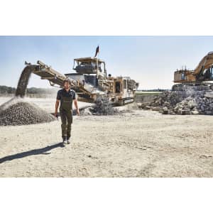 Workwear pants with bib in dark green, worn by a person in a construction setting with machinery and gravel.