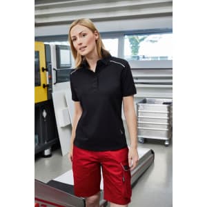 A woman wearing a black polo shirt and red bermuda shorts, standing in an industrial workspace.