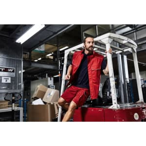 A man wearing a red softshell vest and shorts jumps off a forklift in a warehouse setting. The image includes various boxes.