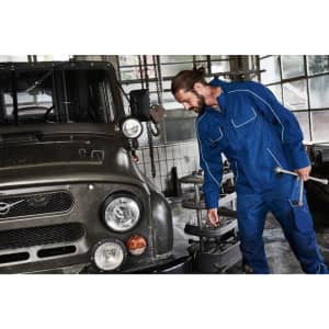 A blue workwear softshell jacket is being worn by a man fixing a vehicle in an industrial setting.