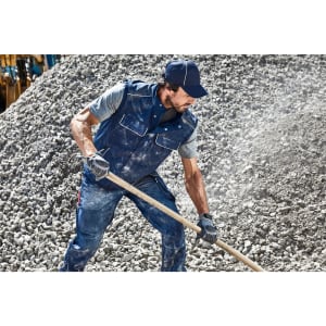 A man wearing a navy blue softshell vest and grey shirt is shovelling gravel. He has gloves and a cap on.