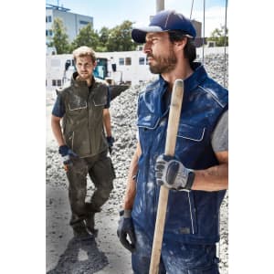 Two men wearing workwear softshell vests, one in navy and one in olive, stand on a construction site.