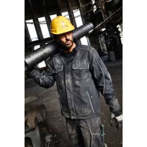 A man in a black softshell jacket carries a pipe over his shoulder, wearing a yellow hard hat and gloves.