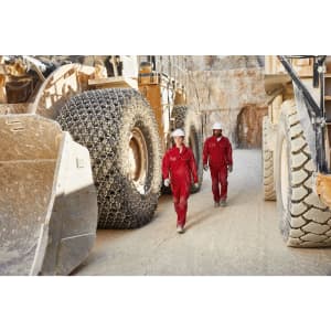 Two workers in red overalls and helmets walk between large machinery in a quarry setting.