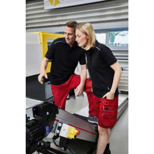 A black men's work polo shirt displayed on two models, paired with red work shorts, in an industrial setting.