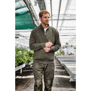 A men's workwear sweat-jacket in olive green, paired with matching work pants, set in a greenhouse environment.