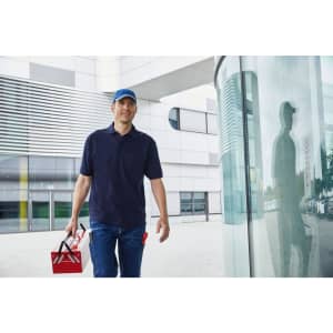 A man wearing a navy polo shirt and cap carries a toolbox while walking past a glass building.