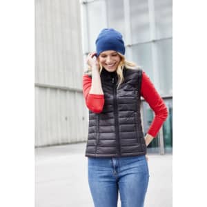 A woman wears a black puffer vest over a red shirt and a blue beanie, smiling outdoors.