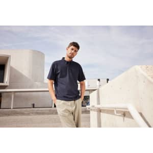 A man wearing a navy plain polo shirt stands casually against a backdrop of modern architecture.