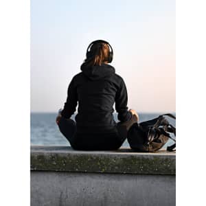 A woman sitting on a ledge wearing a black hoodie and headphones, with a bag beside her, overlooking the water.