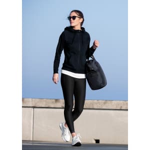 A woman wears a black hoodie and leggings, carrying a bag, walking outdoors against a blue sky.