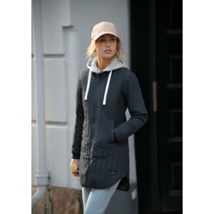 A woman wearing a grey puffer jacket with a hoodie underneath, paired with a beige cap and jeans. The jacket features a logo.