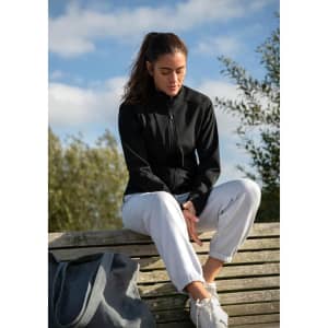 A woman sits on a bench wearing a black soft shell jacket and light trousers, with a bag beside her.
