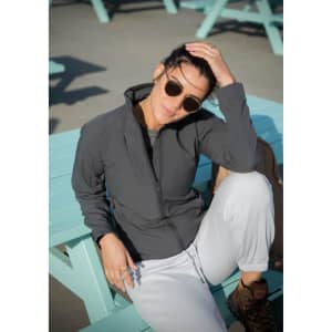 A woman in a grey soft shell jacket sits casually on a light blue table, wearing sunglasses and relaxed attire.