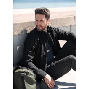 A black soft shell jacket worn by a man sitting on a ledge, with a green bag beside him.