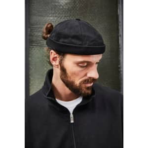 A black chef cap is worn by a man with a beard and a bun, featuring a branded label.