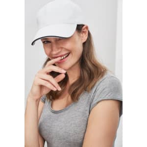 A white six-panel sandwich cap worn by a woman with long hair, smiling and lightly touching her face.