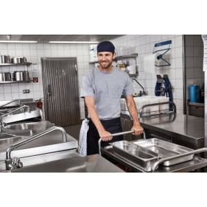 A man wearing a bandana hat pushes a trolley with trays in a commercial kitchen. Stainless steel surfaces are visible.