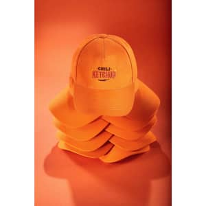 An orange 5-panel cap is displayed on top of a stack of similar caps, featuring a logo on the front.