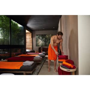 A man wearing an orange bath towel arranges folded towels in a serene spa setting. The room has a natural aesthetic.