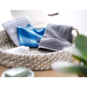 Three hand towels in white, blue, and grey displayed in a woven basket, with a plant and soap nearby.