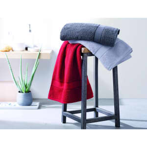 A bath towel display featuring a red, grey, and light grey towel on a black wooden stool, with plants in the background.