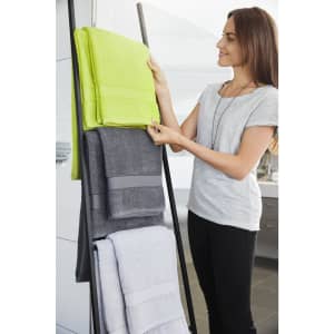 A woman arranges bath towels on a rack, featuring lime green, grey, and white towels. The image includes branding.