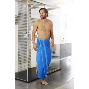 A man wearing a blue bath sheet stands in a modern bathroom near a glass shower.