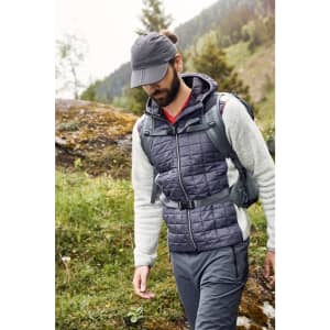 A man wearing a dark grey cap and a quilted vest walks through a green outdoor landscape.