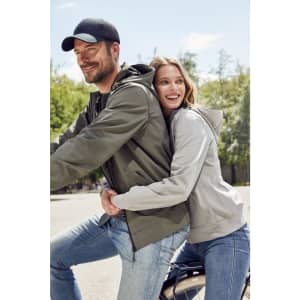 A couple smiles while riding a bicycle together, dressed in casual jackets and a black cap. The scene is set outdoors.