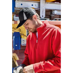 A man wearing a red pullover and black cap works with safety gloves on, surrounded by equipment.