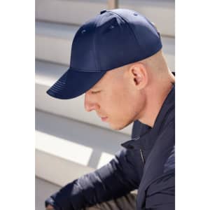 A navy blue 6 panel cap is worn by a man with a shaved head, sitting on stairs. The cap features a logo.