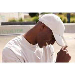 A white 6-panel cap made from bio cotton, worn by a smiling person in a casual setting.