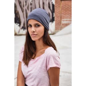A grey Bio Cotton beanie displayed on a model wearing a pink T-shirt, set against an urban backdrop.