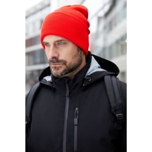 A red high brim beanie worn by a man in a black jacket with a backpack in an outdoor setting.
