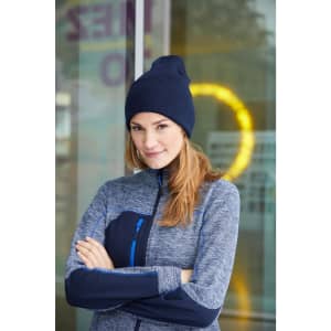 A woman wearing a navy high brim beanie and a grey zip-up jacket stands with her arms crossed. The background features a blurred circle of light.