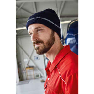 A reflective beanie in navy blue, worn by a man with a beard, inside a hangar or workshop setting.