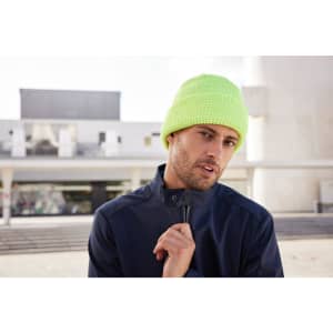 A reflective winter beanie in bright neon yellow, worn by a man in a navy jacket, set against a modern architectural background.