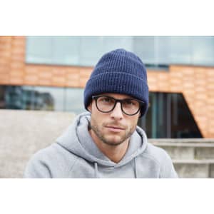 A reflective winter beanie in navy blue, worn by a man in glasses and a grey hoodie, sitting outdoors.
