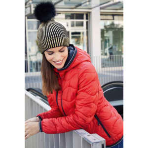 A metallic beanie in gold and black with a pom pom, worn by a woman in a red jacket. Dining area in the background.