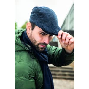 A man in a green jacket and navy scarf tips his blue cap, set against an urban backdrop.