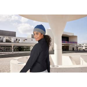 A reversible beanie in blue and grey is worn by a person with curly hair, set against a modern architectural background.