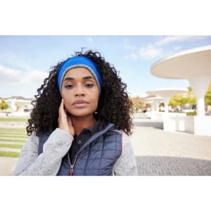 A woman with curly hair is wearing a blue reversible headband and a grey vest in an outdoor setting.