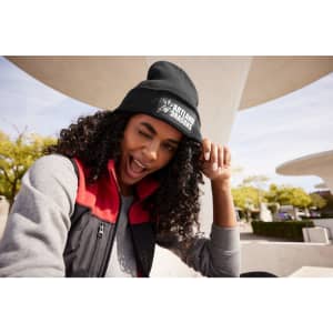 A black knitted beanie with a patch is worn by a person with curly hair, smiling and winking outdoors.