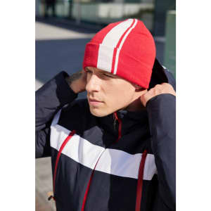 A red winter beanie with white stripes, worn by a person in a navy jacket. The background is an outdoor setting.