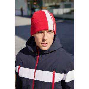 A red winter beanie with white stripes, worn by a person in a navy jacket. The background shows an outdoor setting.
