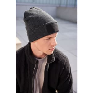 A grey beanie with a patch sits on a model's head, paired with a black jacket in an outdoor setting.